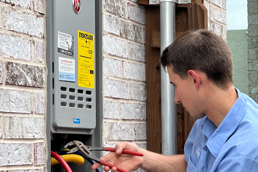 Plumber working on a tankless water heater mounted on the brick exterior wall of a home.